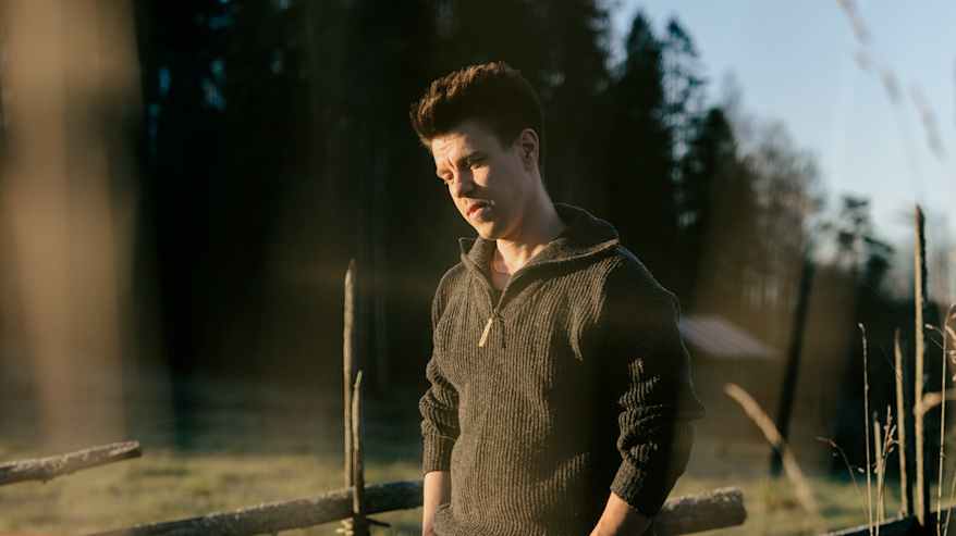 Mies seisoo pellolla syksyisessä maisemassa, taustalla puinen aita ja metsää.