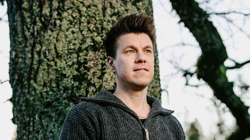 Mies seisoo puun edessä pukeutuneena harmaaseen neulepaitaan ja farkkuihin, pitäen kitaraa käsissään ulkoilmassa.