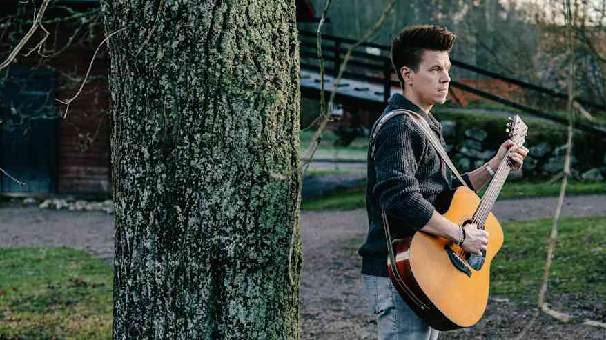 Mies seisoo puun edessä pukeutuneena harmaaseen neulepaitaan ja farkkuihin, pitäen kitaraa käsissään ulkoilmassa.