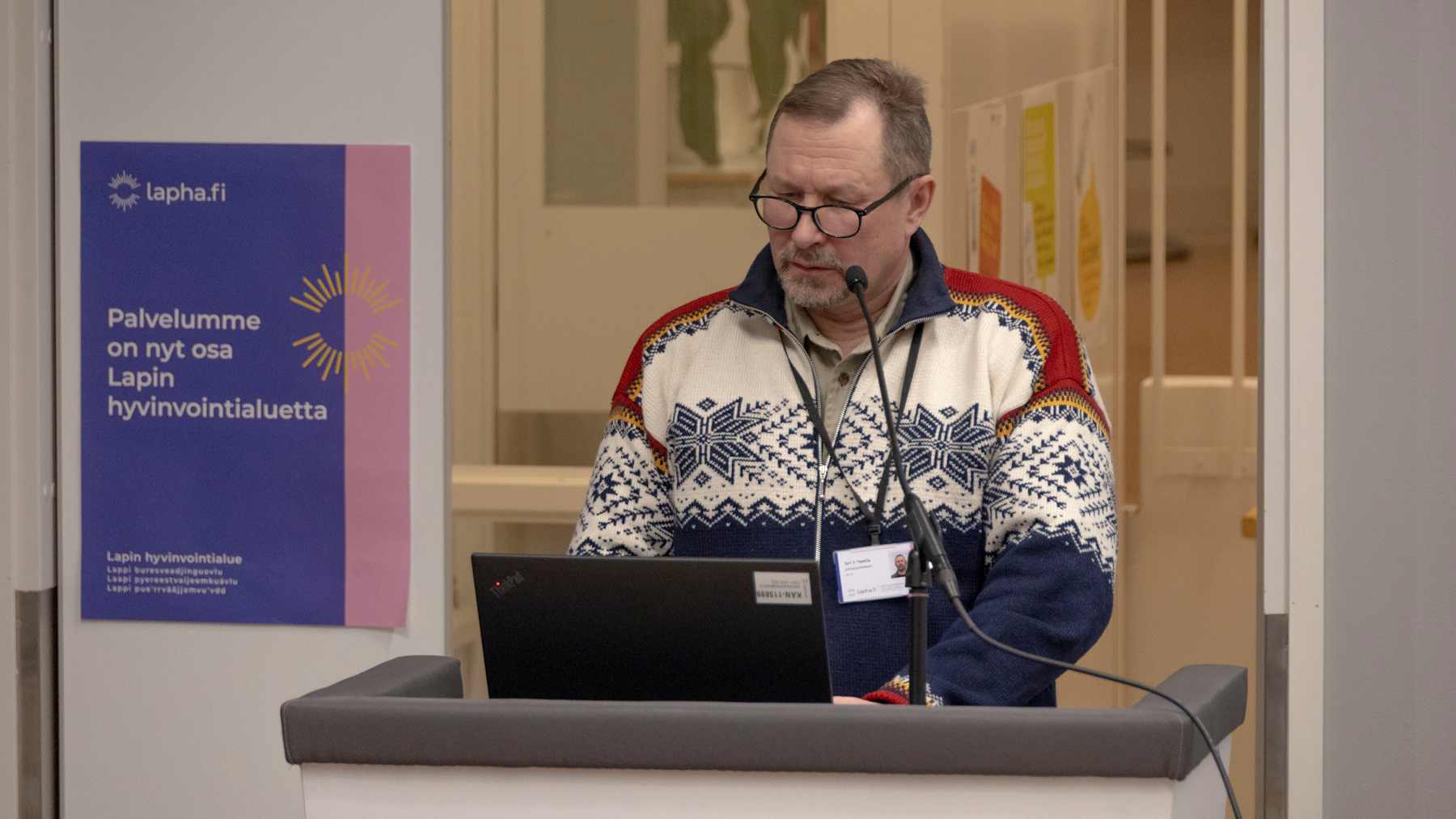 Kuviolliseen villapaitaan pukeutunut mies puhuu mikrofoniin.