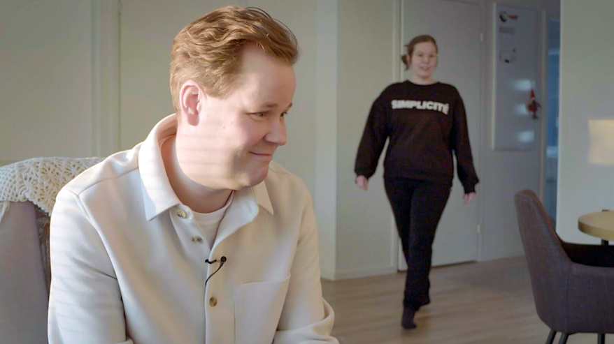 Jussi Laaksonen istuu tuolilla mikrofoni kiinnitettynä hänen paitaansa, ja hänen takanaan kävelee Mari Laaksonen, jonka paidassa lukee "SIMPLICITY"