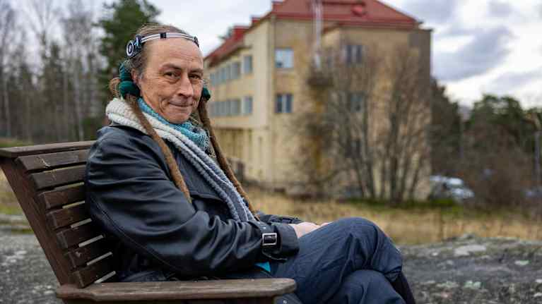 Mies istuu syyspäivänä puutarhatuolissa takanaan keltainen 3 kerroksinen kerrostalo.