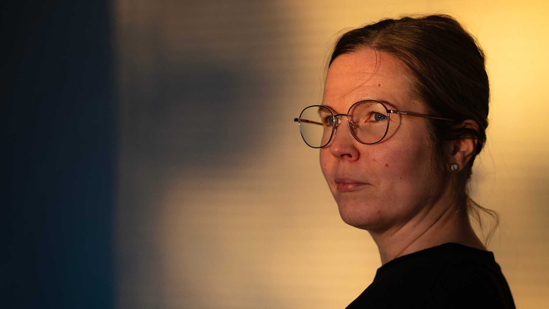 Mari Laaksonen seisoo sivuttain. Hän käyttää silmälaseja sekä mustaa paitaa. Taustalla on lämpimän sävyisiä valoja, jotka luovat varjoja seinälle.