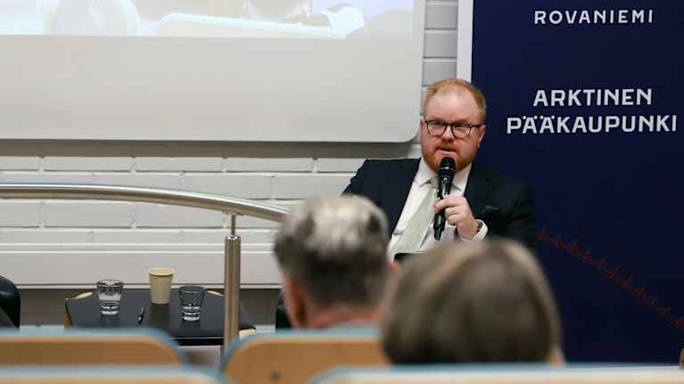 Mies tummassa puvussa istuu puhumassa mikrofoniin.