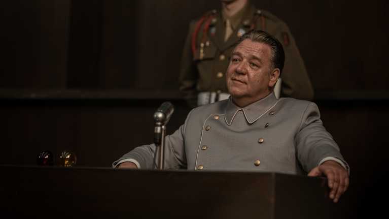 Kuvassa näkyy henkilö virkapuvussa istumassa puupulpetin takana. Pöydällä on mikrofoni. Henkilön taustalla seisoo sotilaspukuun pukeutunut henkilö.