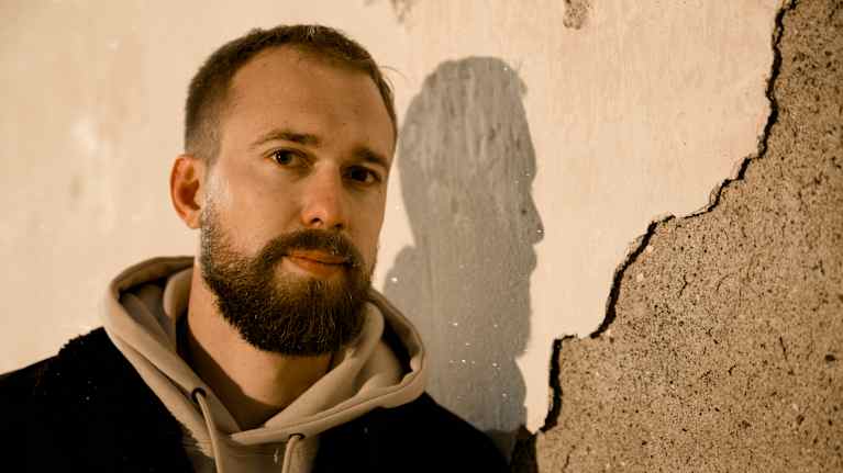 Kuvan henkilö seisoo pinnalta rikkoutuneen betoniseinän edessä pukeutuneena huppariin ja tummaan takkiin.