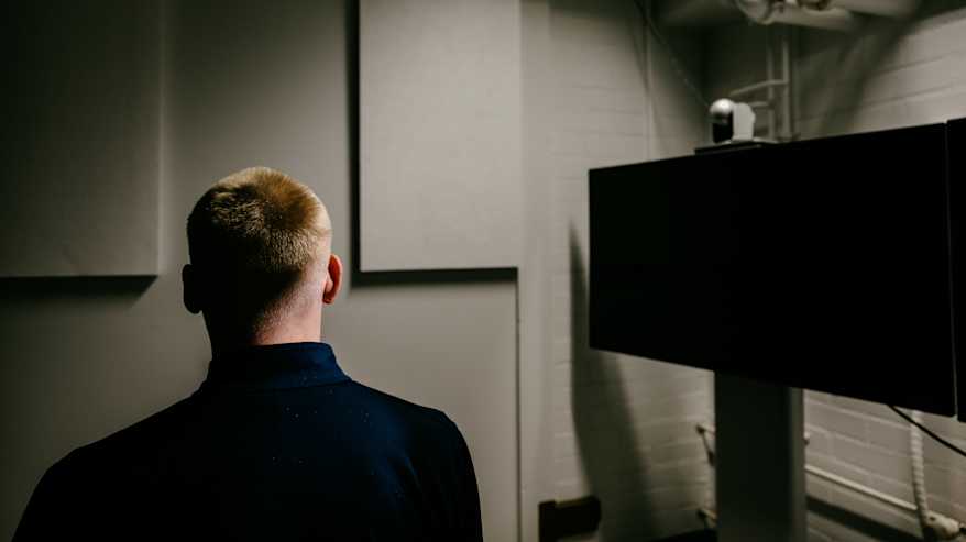 Kuvassa on henkilö selin kameraan hämärässä huoneessa, jossa on monitoreja ja teknisiä laitteita. 