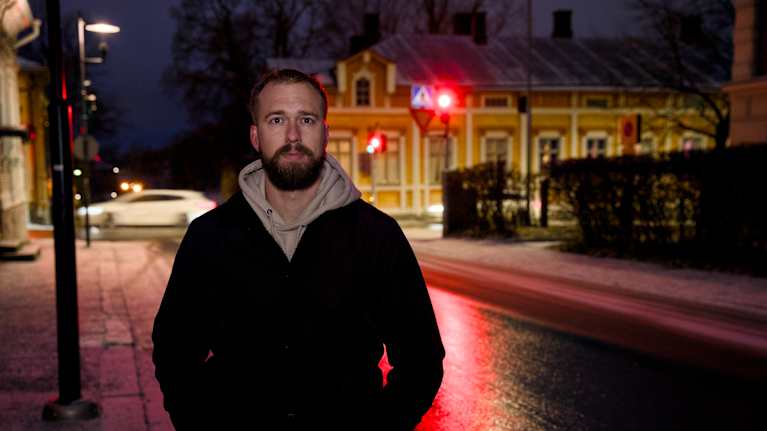  Mies seisoo hämärässä kadun varrella, jossa taustalla näkyy vanha rakennus ja katuun heijastuu punainen liikennevalo.