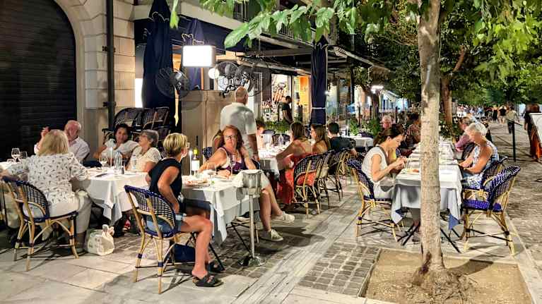 Turisteja istuu kahvilan pöydässä puun varjossa.