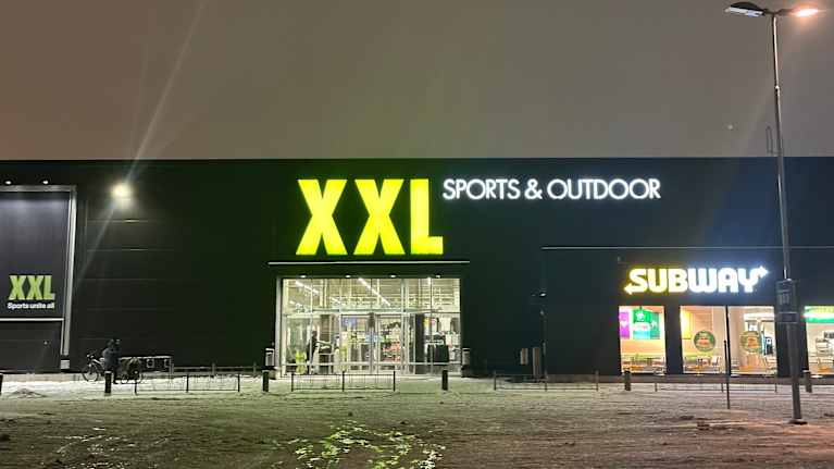 XXL Sports & Outdoor -liikkeen sisäänkäynti iltavalaistuksessa, oikealla Subway-ravintola.