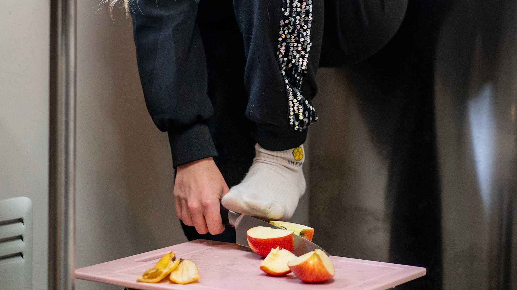 Jenna Laiho leikkaa omenaa pienellä leikkulaudalla käyttäen jalkateräänsä apunaan. Hänen asuaan koristavat kimaltavat yksityiskohdat.