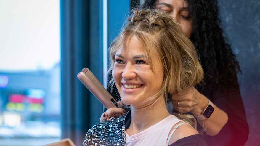 Jenna Laiho hymyilee ja lukee lehteä kampaamossa samalla kun hänen hiuksiaan muotoillaan suoristusraudalla.