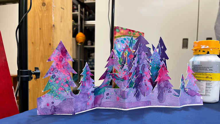 Sinisellä kankaalla on pahvista askarreltu kuusimetsä. Kuuset on väritetty liilan punaisiksi.