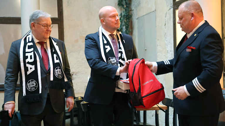 Viking Linen taxfree päällikkö Jamppe Haakana antaa lahjon Viking Linen uudelle toimitusjohtaja Marcus Risbergille. Hänen vieressään seisoo väistyvä toimitusjohtaja Jan Hanses. Heille on annettu harteille jääkiekko TPS:n huivit. Merimiesasuinen Haakana ojentaa punaista laukkua Risbergille.