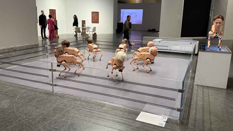 Robottikoiria, joilla on tunnettujen henkilöiden päät näyttelyssä esillä aitiossa. Vieressä korokkeella on robottikoira, jolla Elon Muskin pää.