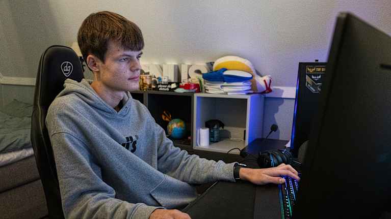 Nuori istuu tietokoneen ääressä huoneessaan ja pelaa videopeliä.