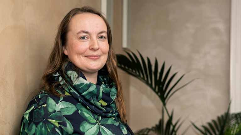 Kuvassa henkilöllä on yllään vihreäsävyinen, lehtikuvioinen huivi ja takki, ja hän seisoo huoneessa, jossa on vaalea seinä ja vehreitä kasveja.