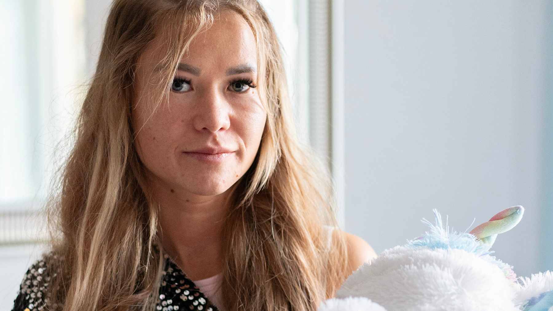 Jenna Laiho istuu sängyllä pitäen sylissään pehmoleluja: pörröinen apina ja valkoinen yksisarvinen. Hänellä on pitkät hiukset ja yllään musta paita.