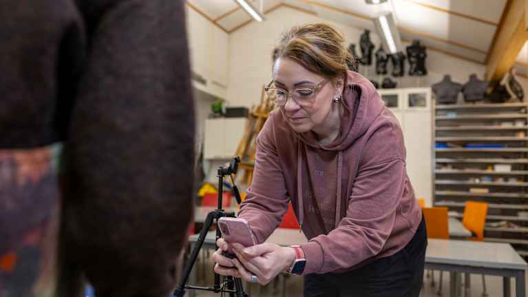 Henkilö kuvaa puhelimella sisätiloissa. Taustalla näkyy hylly ja tuoleja.