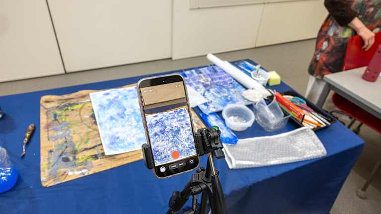 Älypuhelin jalustalla kuvaamassa taidetyöpajan pöydällä olevia materiaaleja. Pöydältä löytyy erilaisia maalaustarvikkeita ja paperiarkkeja.