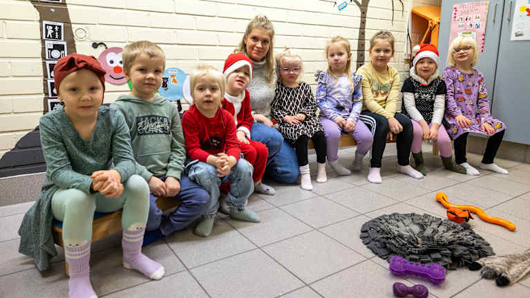 Yhdeksän lasta istuu penkeillä Vehniän päiväkodissa. Lasten keskellä on lastenhoitaja Maija Peura. 