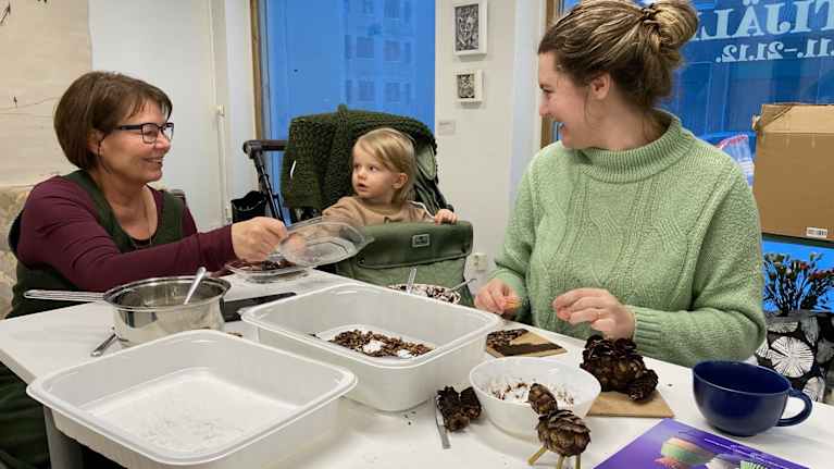 Naiset ja kärryissä istuva taapero askartelevat kävyistä pöydän ääressä.