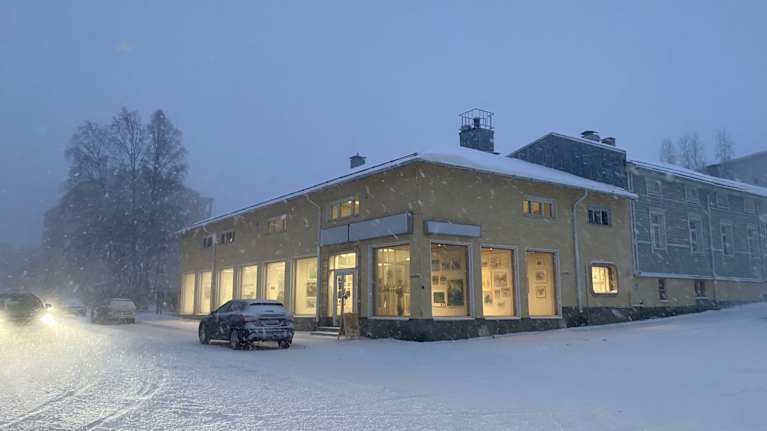 Keltainen kivitalo seisoo katujen kulmauksessa. Suurista näyteikkunoista kajastaa lämmin valo lumipyryn sakeuttamalle kadulle.
