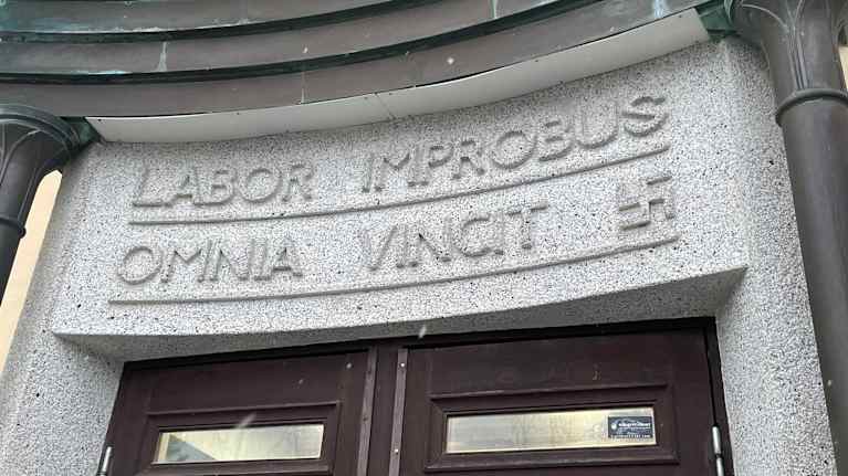 Kuvassa on latinankielinen kyltti koulun oven yläpuolella.