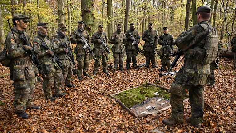 Ryhmä sotilaita seisoo ryhmänjohtajan edessä puolikaaressa syksyisessä metsässä, missä maanpinta on täynnä pudonneita lehtiä.