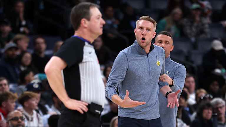 Koripallovalmentaja Tuomas Iisalo huutaa kentän laidalla ottelun erotuomarille, Iisalon avustaja pitää häntä käsivarresta kiinni.