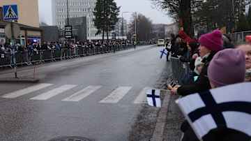 Ihmisiä odottaa kadun varrella, ja joukossa näkyy useita Suomen lippuja. Taustalla poliisiauto.