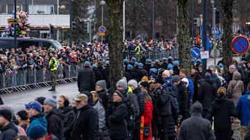 Suuri joukko ihmisiä kadun molemmin puolin. Taustalla kasa folioilmapalloja. Kadulla marssii sinibarettisia tummiin pukeutuneita ihmisiä.