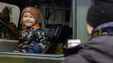 Hymyilevä lapsi Viljo Ruoppa istuu sotilasajoneuvon ohjaamossa, kun aikuinen tarkkailee tilannetta ulkopuolelta.