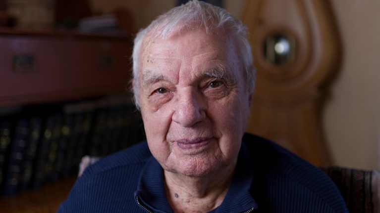 Vanha mies katsoo suoraan kameraan. Hänellä on sininen paita päällä.