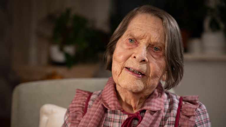 Vanha nainen on pukeutunut punaruudulliseen paitaan. Hänen päänsä on hieman vinossa.