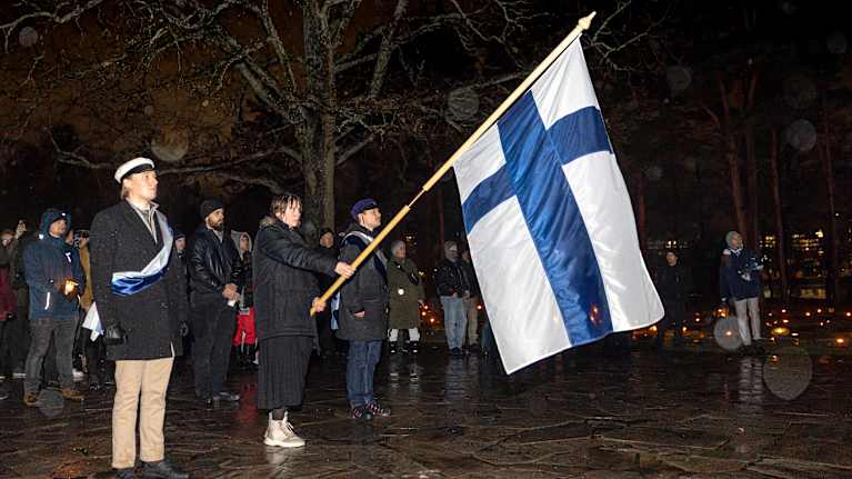 Nainen pitää suurta Suomenlippua. Vieressä seisoo mies valkoinen ylioppilashattu päässä.