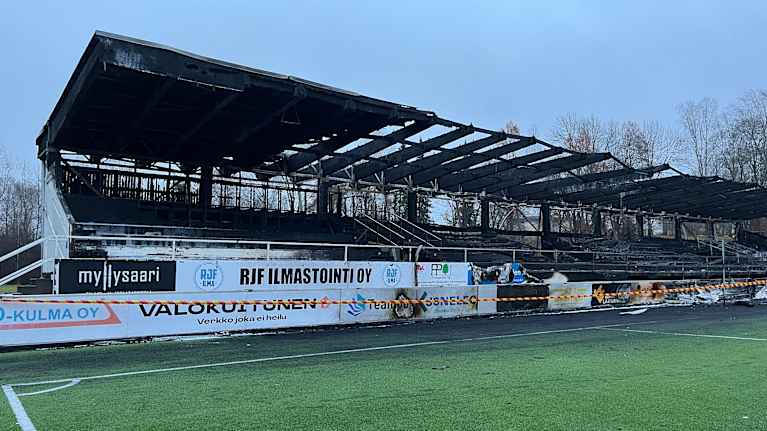 Tulipalossa tuhoutunut jalkapallokatsomo, jonka edessä eritysnauha. Etualalla jalkapallokentän nurmea.