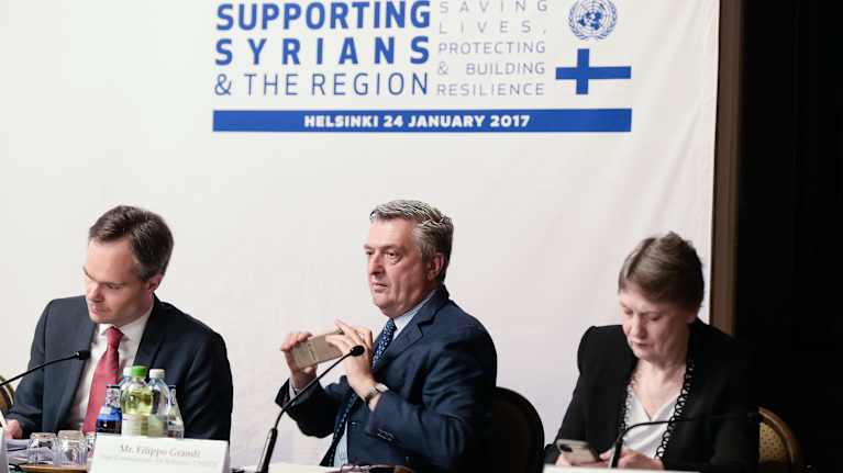 Kolme henkilöä istuu pöydän ääressä, taustalla kyltissä lukee Supporting Syrians and the region.