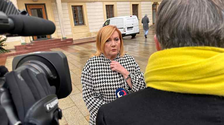 Vaalea myöhäiskeski-ikäinen nainen mustavalkoruutuisessa takissa kameroiden ja mikkien edessä kivisellä, märällä pihalla.