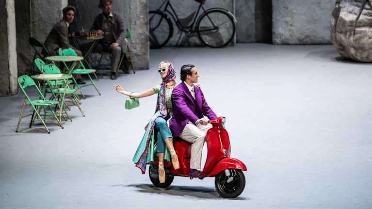 Mies ajaa punaisella vespalla, kyydissään nainen, jolla on balettitossut ja aurinkolasit.