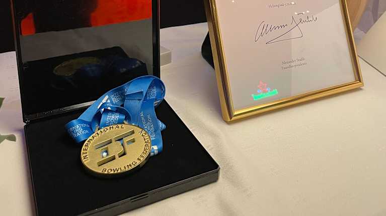 Luukas Väänäsen keilailun miesten sarjan MM-kultamitali, lajiliiton viiri, presidentti Stubbin onnittelukirje ja keila juhlapöydällä keilahallissa.