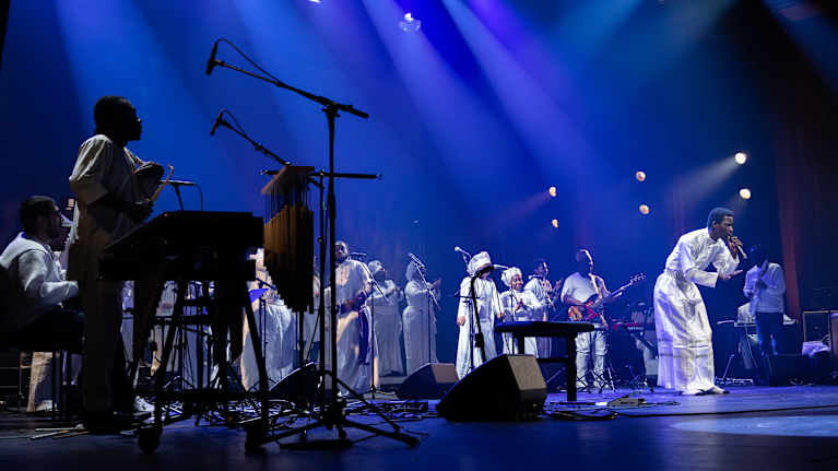 Kuvassa on energinen esiintyminen lavalla, jossa useat muusikot ovat pukeutuneet valkoisiin asusteisiin. 