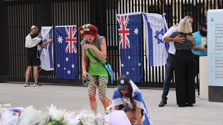 Aitaan on kiinnitetty Australian ja Israelin lippuja. Kaksi ihmistä on kumartunut maassa olevien kukkien puoleen. Taustalla kaksi ihmistä halaa. 