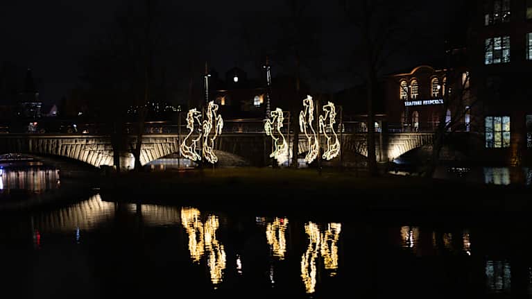 Kuvassa on yöaikaan valaistu joki, jonka varrella on valoteoksia. Sillat ja rakennukset näkyvät taustalla, ja valot heijastuvat veden pinnasta.