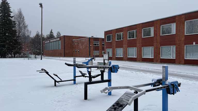 Etualalla on ulkokuntosalin välineitä ja taka-alalla kaksi isoa punatiilirakennusta. Luminen maa.