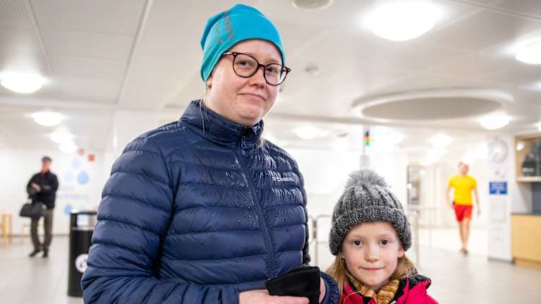 Kuvassa nainen ja tyttö ulkovaatteet päällä uimahallin aulassa.