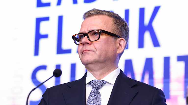 Petteri Orpo seisoo mikrofonin edessä puvussa.