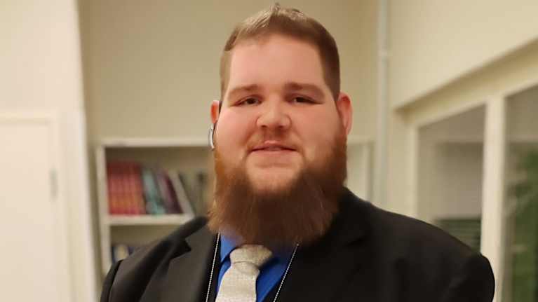 Parrakas mies mustassa puvussa katsoo kameraan.