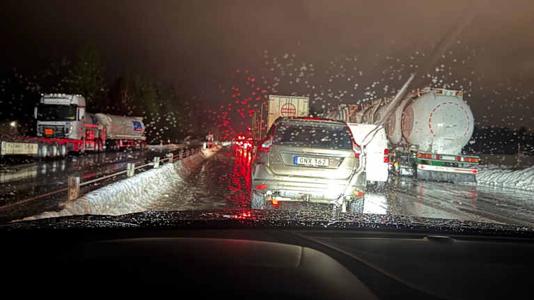 Pitkä autojono pimeällä tiellä, etualalla kolme rekkaa. Tie on kiiltävä ja jäinen. Auton ikkunassa on runsaasti vesipisaroita.