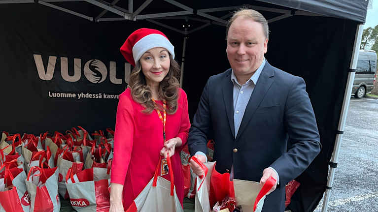 Lukuisia punaisia ja harmaita lahjakasseja rivissä täynnä elintarvikkeita, kaksi ihmistä pitää kasseja käsissään.